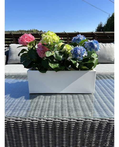 Den Kunstige Plante Hortensia Pink UV 35 cm, en rektangulær hvid plantekasse med lyserøde hortensiaer, er placeret udenfor på et fletbord med en glasplade, grå hynder og en blå himmel i baggrunden.