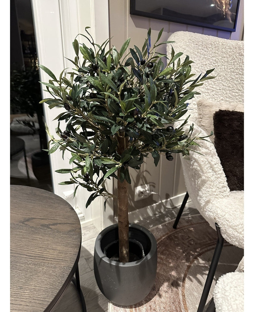 Det kunstige oliventræ UV 85 cm, en kompakt og letplejet kunstig plante med mørkegrønne blade og sorte oliven, står indendørs ved siden af et rundt træbord og en lænestol med hvid tekstur.