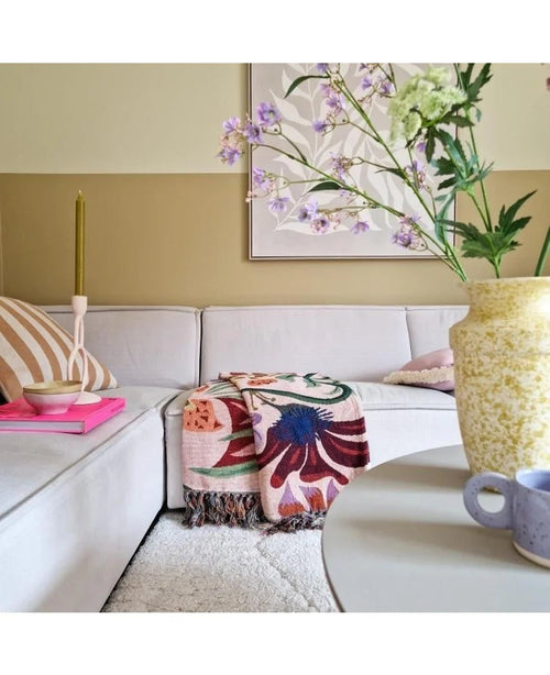 En hyggelig stue med en lys beige sofa, et farverigt tæppe med blomster og mønstrede puder. På et rundt bord står en gul vase med Kunsttak Daphne Spray 108 cm lilla og en lille blå kop. Kunst hænger på den neutrale væg.