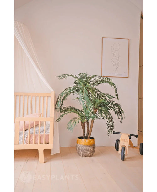 Et hyggeligt børneværelse med lyst træseng, Artificial Palm Cycas Classic 135 cm i en gul plantekasse, en legetøjscykel i træ med mærket MIA, minimalistisk streg på væggen, lyse trægulve og blødt pink sengetøj for ekstra varme.