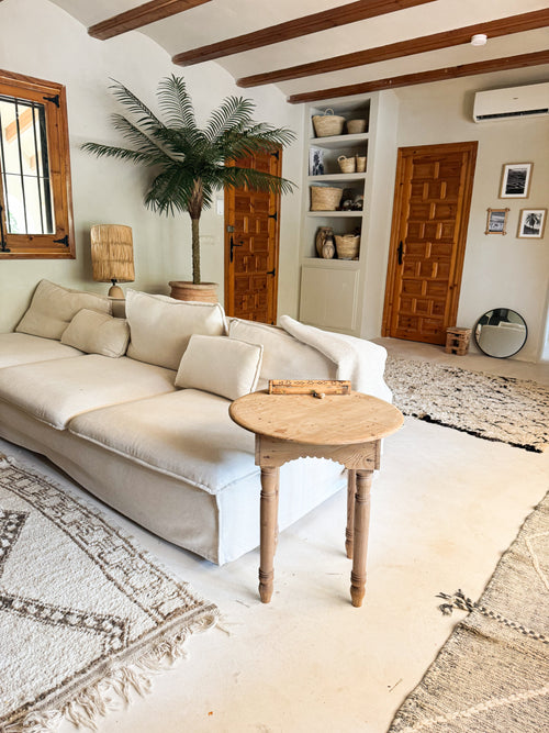 En hyggelig stue med hvid sofa, rundt sidebord i træ, kunstig palme Phoenix De Luxe 210 cm, vævede kurve på hylder, trælåger, et rundt spejl og neutralt tonede tæpper på et lyst gulv.