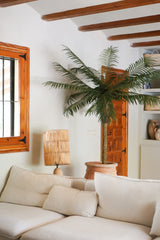 En hyggelig stue med en hvid sofa, en rattan bordlampe, Artificial Palm Phoenix De Luxe 210 cm i en potte, træbjælker i loftet, et vindue med trærammer og en trædør i baggrunden.