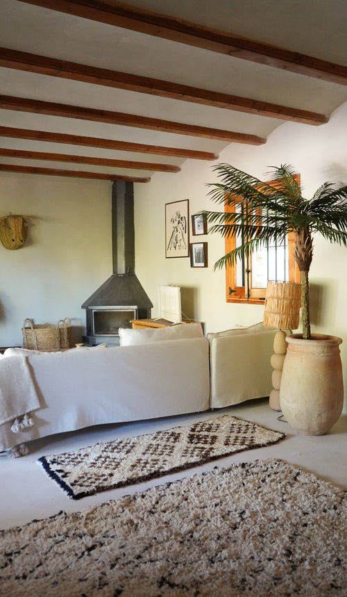 En hyggelig stue har hvid sofa, mønstret tæppe, brændeovn, indrammet kunst og træbjælker. Sollyset strømmer ind fra et vindue ved siden af den frodige Artificial Palm Phoenix De Luxe 210 cm og vækker rummet til live.