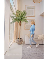En kvinde i blå bluse og jeans går forbi Kunstpalm Phoenix De Luxe 250 cm i en lys, moderne stue med høje vinduer, neutral indretning og indrammet kunst på væggen.