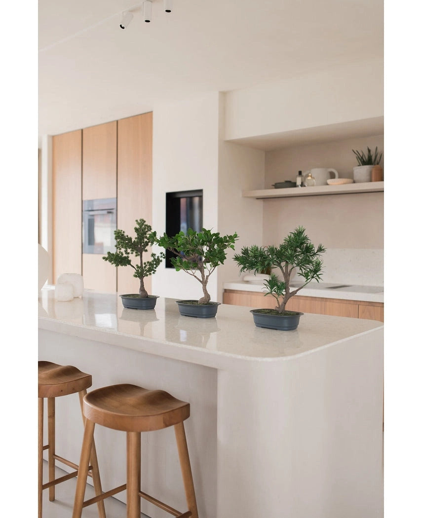 Een Kunstplant Bonsai Juniper 40 cm staat op een wit keukeneiland met twee houten krukjes, omringd door lichthouten kastjes, witte werkbladen en een minimalistische inrichting.