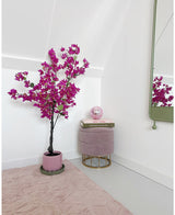 Et moderne, minimalistisk værelseshjørne med en kunstig bougainvillea 125 cm Lilla plante i en krukke, en rund pink fodskammel med guldben, stablede bøger, en pink krystalkugle, et vægspejl og et blødt pink tæppe på et lyst gulv.