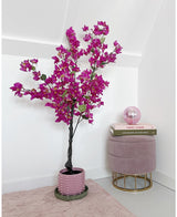 Den kunstige bougainvillea 125 cm Lilla med levende lilla blomster står ved siden af en pink fløjlsskammel med bøger stablet ovenpå og en pink glaskugle i et minimalistisk, moderne rum med hvide vægge.