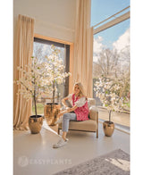 En kvinde griner på en beige lænestol nær store vinduer med beige gardiner, omgivet af tre kunstige bougainvillea 150 cm hvid i et lyst, moderne opholdsrum fuld af naturligt lys.