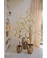 En kunstig bougainvillea-plante, der måler 150 cm høj, står i en dekorativ guldkrukke nær et solbelyst vindue med beige gardiner og tilføjer elegance til det hyggelige rum. Nærliggende bogreoler fremhæver den indbydende interiør atmosfære.