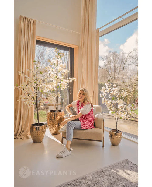 En kvinde i en pink mønstret cardigan og jeans sidder på en beige lænestol nær store vinduer med cremefarvede gardiner, omgivet af tre 180 cm hvide kunstige bougainvillea-planter i skinnende guldplantekasser. Sollys fylder det moderne, luftige rum.