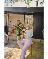 En blond kvinde i hvid bluse og lyse lilla bukser sidder udenfor på en skammel med den kunstige Calathea Makoyana UV 75 cm rød potteplante, på et træterrasse foran et moderne hus med glasdøre.