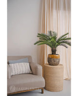 En Artificial Cycas Palm 80 cm står på et rundt fletbord ved siden af en beige lænestol med en hvid mønstret pude, mod lyse beige gardiner og en neutral væg.