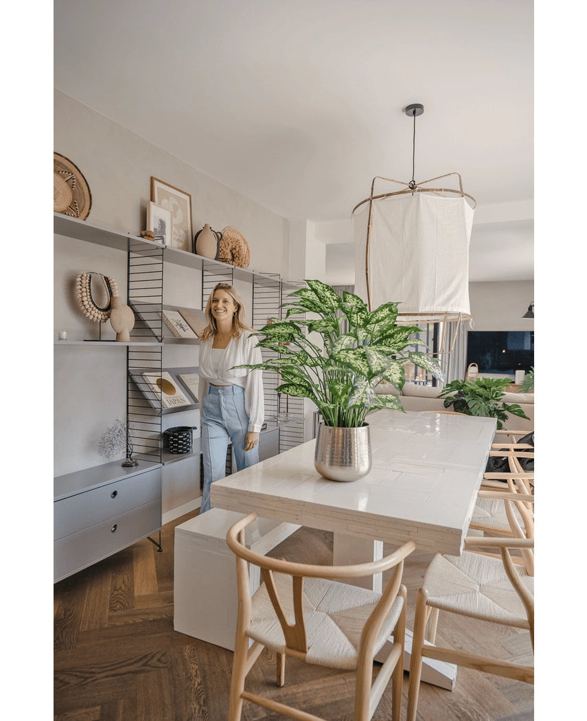 Een vrouw glimlacht in een lichte, moderne eetkamer met houten stoelen, een witte tafel met de Kunstplant Dieffenbachia 75 cm als middelpunt en open planken versierd met boeken en kunst.