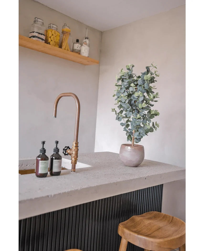 En moderne bordplade har en guldhane, tre sæbedispensere, den kunstige eukalyptus 91 cm plante og træskamler. Minimalistiske hylder over viser krukker med forskellige ingredienser for et hyggeligt look.