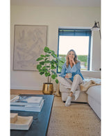 En kvinde sidder på en beige sofa i en hyggelig moderne stue, holder et krus og smiler. Rummet er præget af den kunstige ficusplante 120 cm i en guldkrukke, abstrakte kunstværker og sollys, der skinner gennem blå persienner.