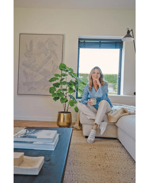 En kvinde sidder på en beige sofa i en hyggelig moderne stue, holder et krus og smiler. Rummet er præget af den kunstige ficusplante 120 cm i en guldkrukke, abstrakte kunstværker og sollys, der skinner gennem blå persienner.