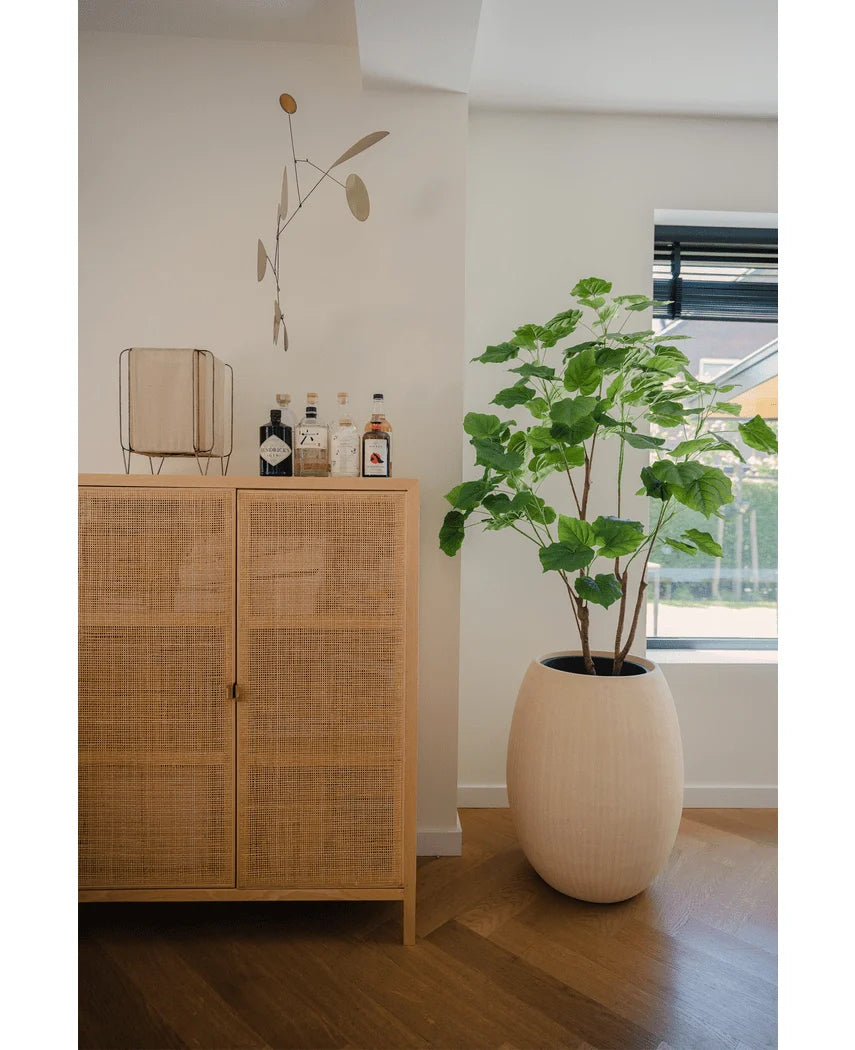 Et lyst træskab med flasker og indretning står ved siden af en kunstig ficus plante 150 cm i en cremefarvet plantekasse nær et vindue med persienner, i et lyst moderne rum med trægulv.