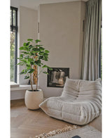 En hyggelig, beige loungestol med struktur står ved siden af Artificial Ficus planten 150 cm i en moderne stue med lyse trægulve, neutrale vægge, et vindue og gulvlange grå gardiner.