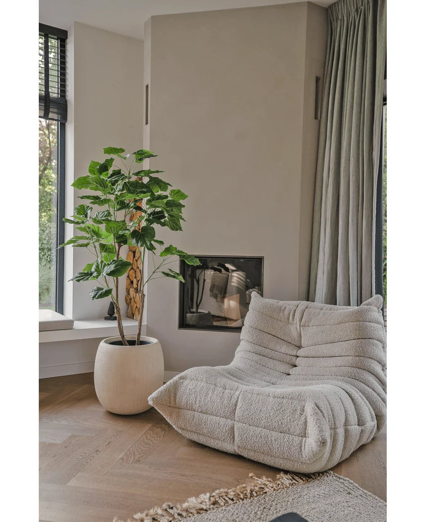 En hyggelig, beige loungestol med struktur står ved siden af Artificial Ficus planten 150 cm i en moderne stue med lyse trægulve, neutrale vægge, et vindue og gulvlange grå gardiner.