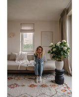 En kvinde med krøllet blond hår sidder på en beige sofa i en lys stue og hviler hagen på hænderne. Ved siden af hende står UV-planten Artificial Hydrangea 85 cm høj, oplyst af lys gennem et vindue med beige gardiner.
