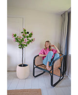 En kvinde i lyserød sweater og blå jeans sidder barfodet på en rattanstol ved siden af den kunstige plante Judas 150 cm, et kunstigt træ med lyserøde blomster, i et lyst, moderne rum med hvide vægge og gulve.