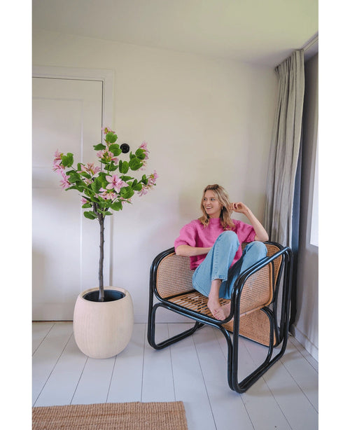 En kvinde i lyserød sweater og blå jeans sidder barfodet på en rattanstol ved siden af den kunstige plante Judas 150 cm, et kunstigt træ med lyserøde blomster, i et lyst, moderne rum med hvide vægge og gulve.