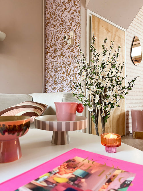 En hyggelig stue med en pink og pastelfarvet indretning byder på en stribet bakke til fods, en pink kop, et lys, en skål, et livligt magasin og den kunstige kirsebærblomstplante 120 cm creme i en høj vase nær en mønstret væg og trædør.