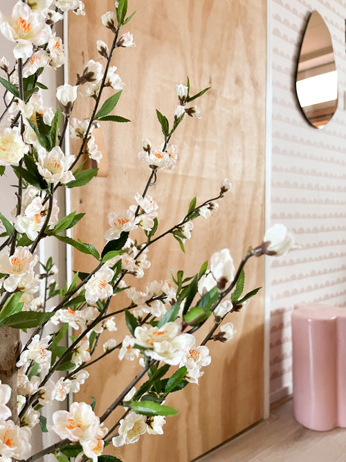 En kunstig kirsebærblomstplante 120 cm creme med hvide og lyserøde blomster står foran en trædør. Bag den er en pink skammel, spejl og pink mønstret tapet.