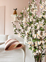 En hyggelig stue med en hvid sofa, en stribet pink og brun pude og den kunstige kirsebærblomst 120 cm cremeblomstrende gren i forgrunden. En potteplante hviler på en væghylde mod en lyserød væg.