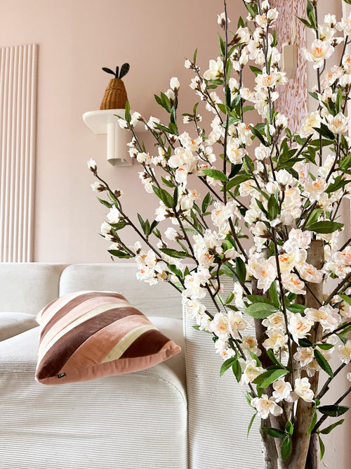 En hyggelig stue med en hvid sofa, en stribet pink og brun pude og den kunstige kirsebærblomst 120 cm cremeblomstrende gren i forgrunden. En potteplante hviler på en væghylde mod en lyserød væg.
