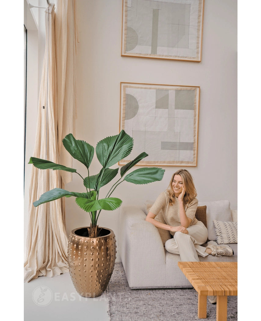Een vrouw ontspant op een lichte bank in een moderne, neutraal gekleurde woonkamer. Naast haar staat de Kunstplant Lucuala Grandis 120 cm in een pot, terwijl er abstracte kunst boven hangt en natuurlijk licht de ruimte verlicht.