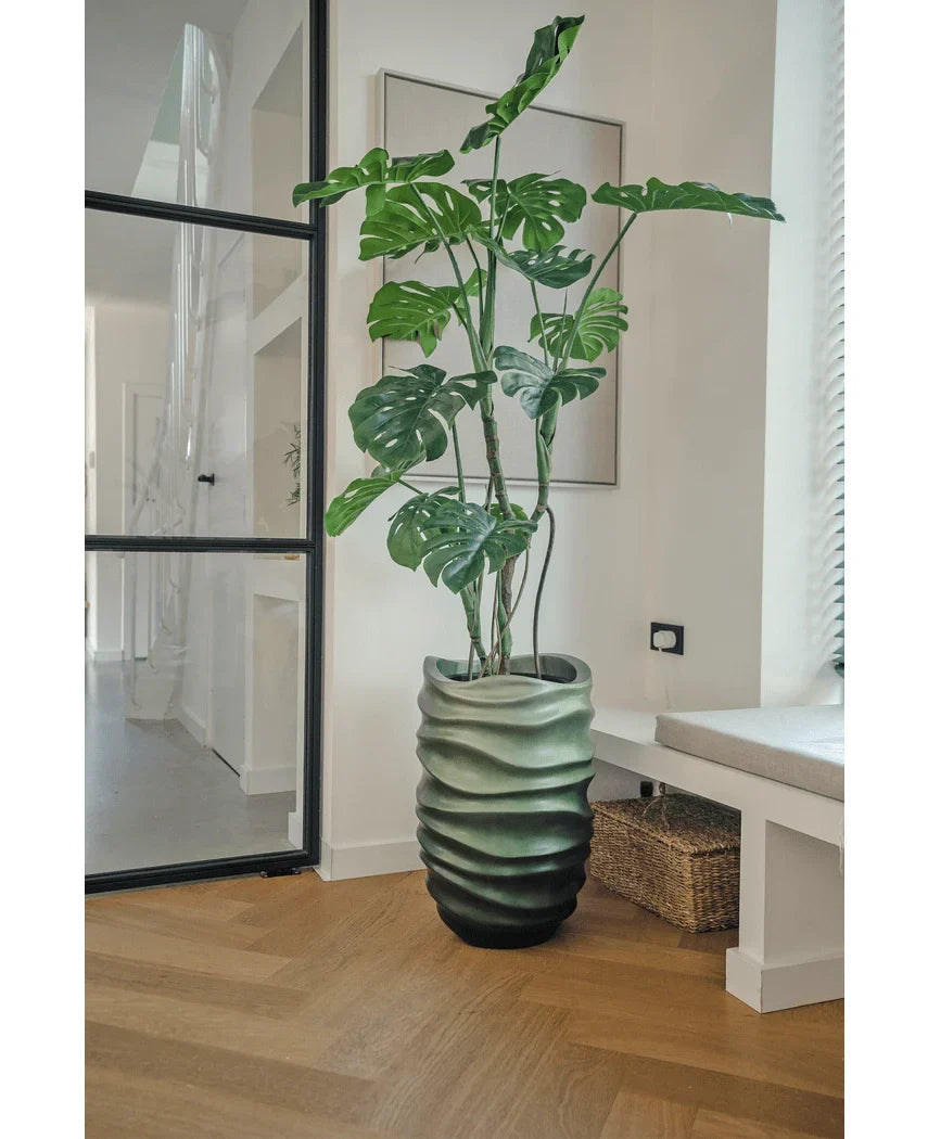 Kunstplanten Monstera 150 cm, med sine store grønne blade, står i en krukke med bølget mønster på et trægulv i et moderne, lyst rum med glasdør, indrammet kunst og en flettet kurv i nærheden.