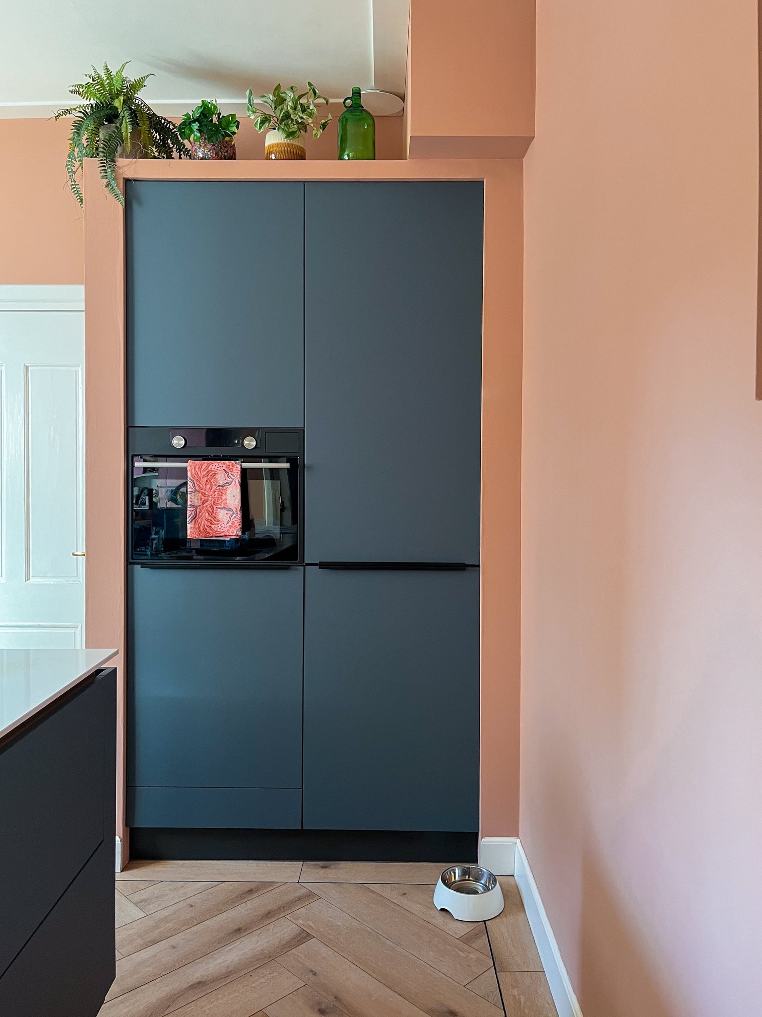 Een moderne keukenhoek met matte donkere kasten, een ingebouwde oven en een lichte houten vloer. De Kunstplant Monstera 18 cm en een groene glazen fles staan bovenop de kastjes; een voerbak voor huisdieren staat op de perzikkleurige muur.