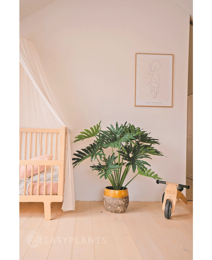 Een gezellige, minimalistische kinderkamer met links een houten wieg en luifel, in het midden een Kunstplant Philodendron UV 90 cm als pronkstuk, rechts een houten speelgoedfiets en daarboven een eenvoudige lijnkunstprint.