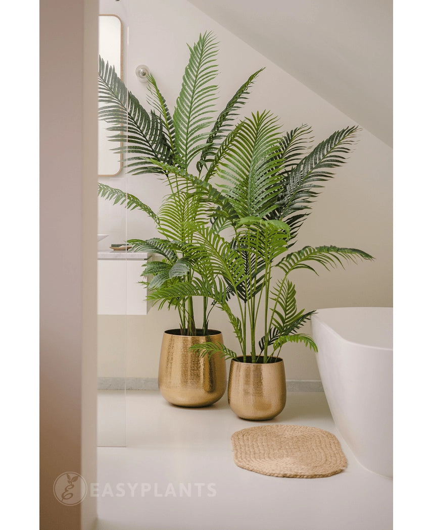 Twee Kunstplant Phoenix 160 cm met lange, groene bladeren in glanzende gouden potten geven een luxe touch aan de witte badkamervloer bij het bad en de ronde geweven mat. Op de achtergrond zijn een spiegel en een wit aanrecht te zien.