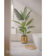 Kunstplanten Phoenix 180 cm i guldkrukke står ved siden af et hvidt badekar og håndvask i et moderne minimalistisk badeværelse med hvide vægge og gulv. I nærheden ligger en rundvævet måtte på gulvet.