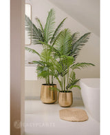 En kunstig plante Phoenix 180 cm med frodige grønne blade i en skinnende guldkrukke står ved siden af et hvidt badekar og en beige vævet måtte i et moderne, minimalistisk badeværelse, suppleret med et spejl og et vindue for et fredfyldt look.