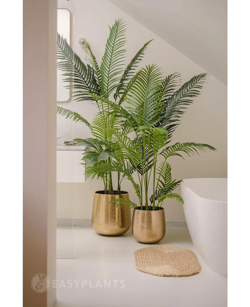 En kunstig plante Phoenix 180 cm med frodige grønne blade i en skinnende guldkrukke står ved siden af et hvidt badekar og en beige vævet måtte i et moderne, minimalistisk badeværelse, suppleret med et spejl og et vindue for et fredfyldt look.