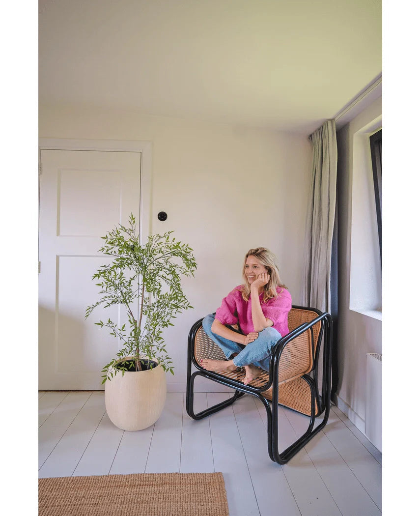 En kvinde i lyserød skjorte og jeans sidder barfodet på en vævet stol og smiler og slapper af i et lyst, minimalistisk rum med hvide vægge og gulve. Ved siden af hende står Artificial Ruscus 120 cm-planten.