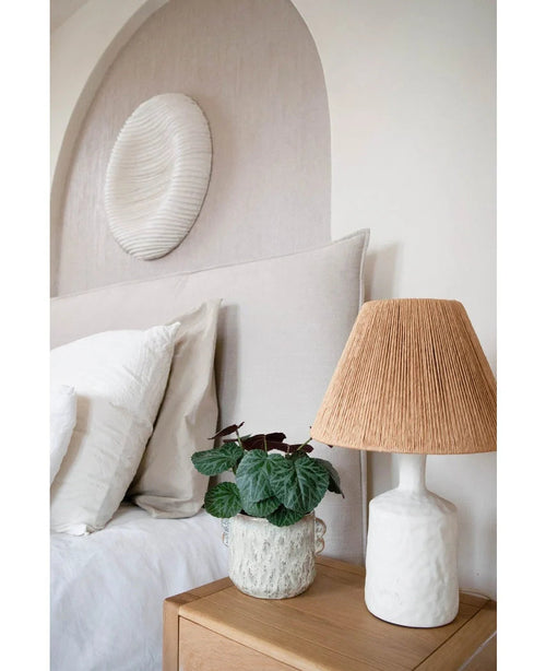 Et moderne soveværelse med en neutral farvepalet har en seng med hvidt sengetøj, et træ sengebord, en struktureret keramisk lampe med vævet skærm, den Artificial Saxifraga plante 27 cm og en rund hvid vægskulptur over sengegavlen.