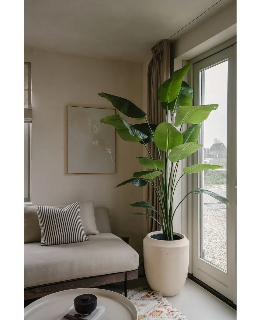 De Kunstplant Strelitzia 180 cm real touch staat bij de glazen deuren in een lichte, moderne woonkamer met een beige bank, gestreept kussen, minimale muurkunst en een neutrale inrichting.