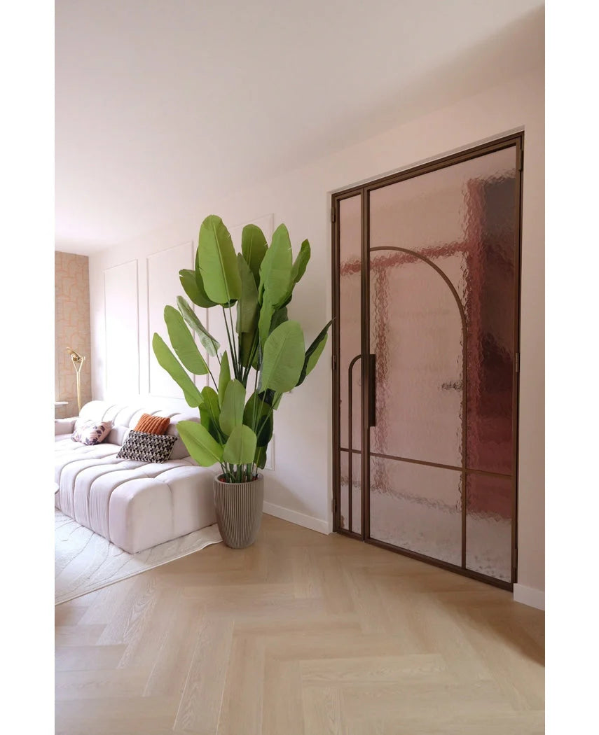 En moderne stue med hvid sofa, mønstrede hynder og kunstplanten Strelitzia Real Touch Deluxe 210 cm. Et lyst trægulv, bløde lyserøde vægge og en glasdør med bronzemetalramme skaber en let og luftig atmosfære.