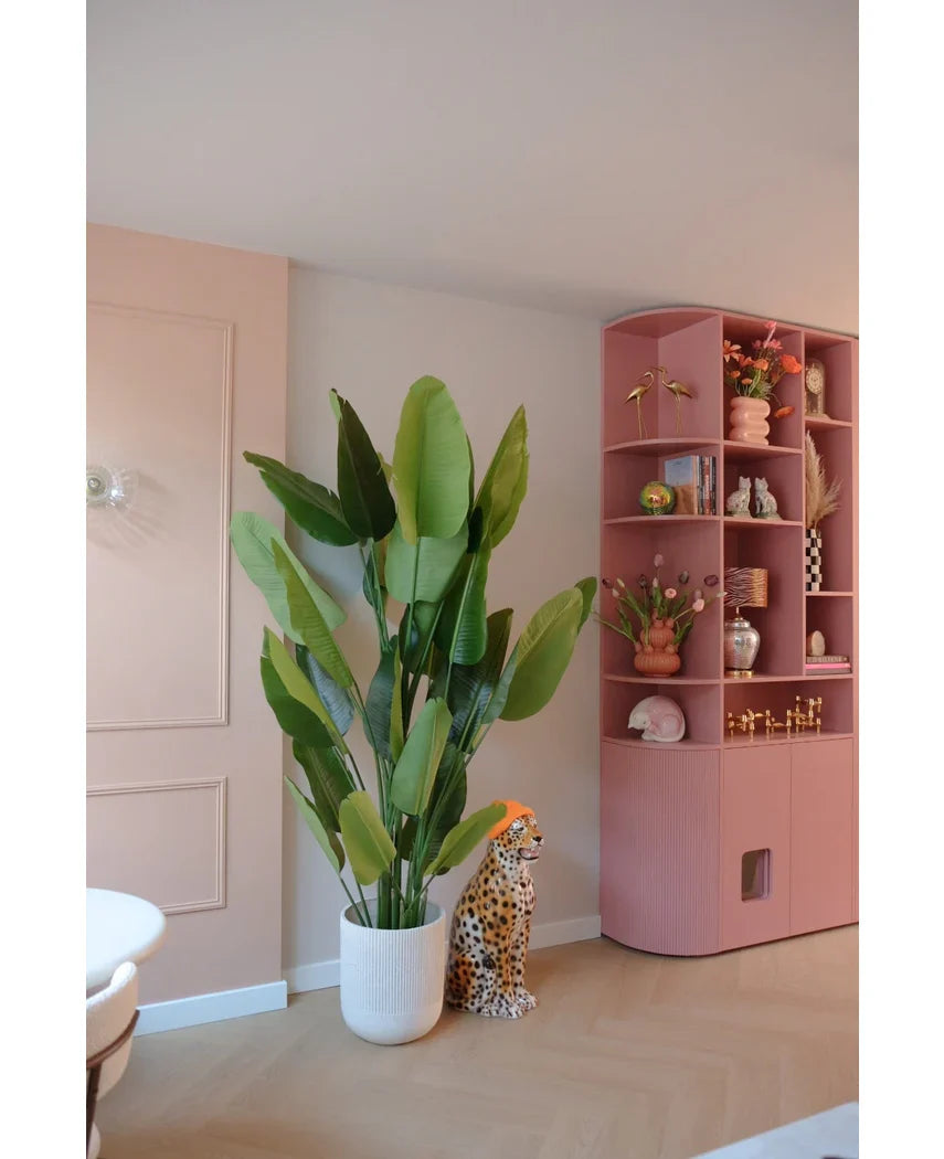 De Kunstplant Strelitzia Real Touch Deluxe 210 cm staat bij een cheetafiguur van keramiek met een oranje hoed, naast een hoge roze plank vol vazen, bloemen en ornamenten in een pastelkleurige kamer.