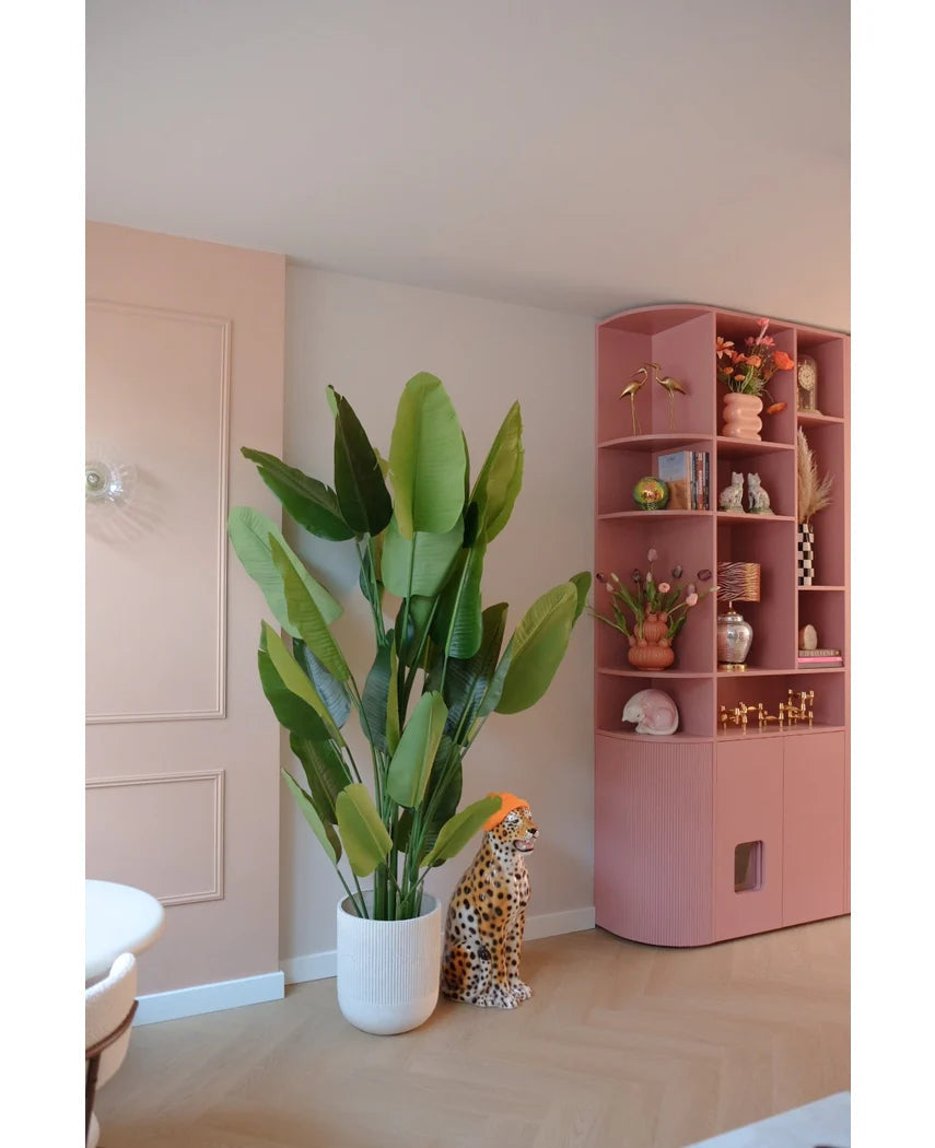 Den kunstige plante Strelitzia Real Touch Deluxe 210 cm står ved siden af en keramisk gepardfigur med en orange hat, ved siden af en høj pink hylde fuld af vaser, blomster og ornamenter i et pastelfarvet rum.