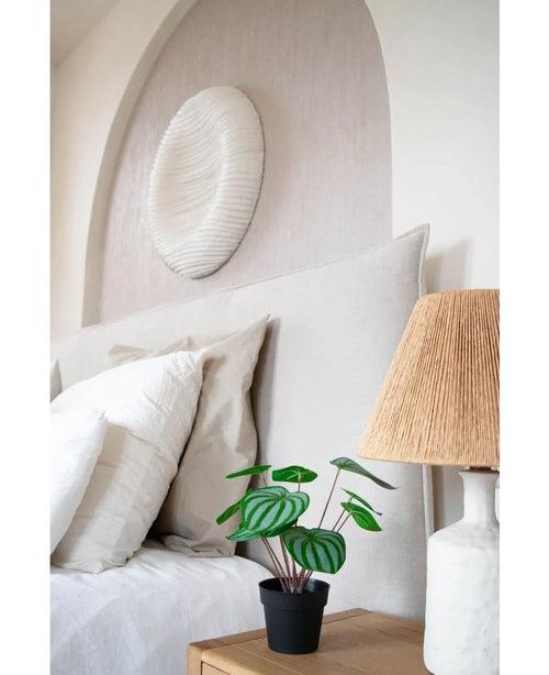 En pænt lavet seng med hvide puder og sengetøj står ved siden af et træ sengebord med den kunstige vandmelonplante 29 cm og en lampe med vævet skærm; over sengegavlen hænger en hvid tekstureret vægskulptur.