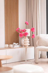 En moderne stue har et hvidt sidebord med Artificial Branch Cherry Blossom 85 cm pink, hvortil der er tilføjet lyse lyserøde blomster. Neutral indretning, lyse træaccenter, beige gardiner og en struktureret lænestol fuldender det hyggelige, minimalistiske rum.