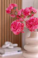 En hvid keramikvase rummer en kunstig Kirsebærblomstgren 85 cm pink. Baggrunden viser lodrette træpaneler, en stak bøger og en lille dekorativ genstand på toppen.