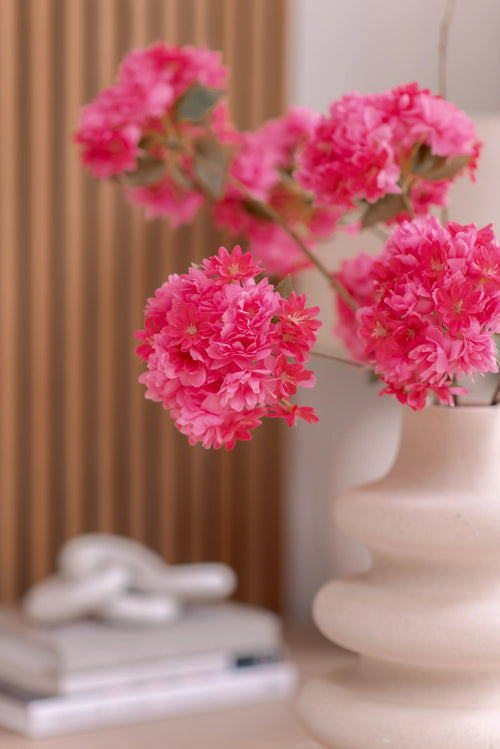 En hvid keramikvase rummer en kunstig Kirsebærblomstgren 85 cm pink. Baggrunden viser lodrette træpaneler, en stak bøger og en lille dekorativ genstand på toppen.