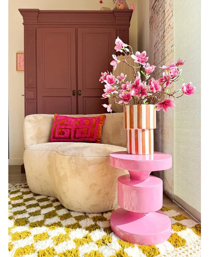 En hyggelig stue med en beige rund sofa, en pink og orange geometrisk pude, et pink sidebord, kunstig gren Magnolia lille blomstret 89 cm pink i en vase, et gul-hvidt mønstret tæppe og et kastanjebrunt skab i baggrunden.