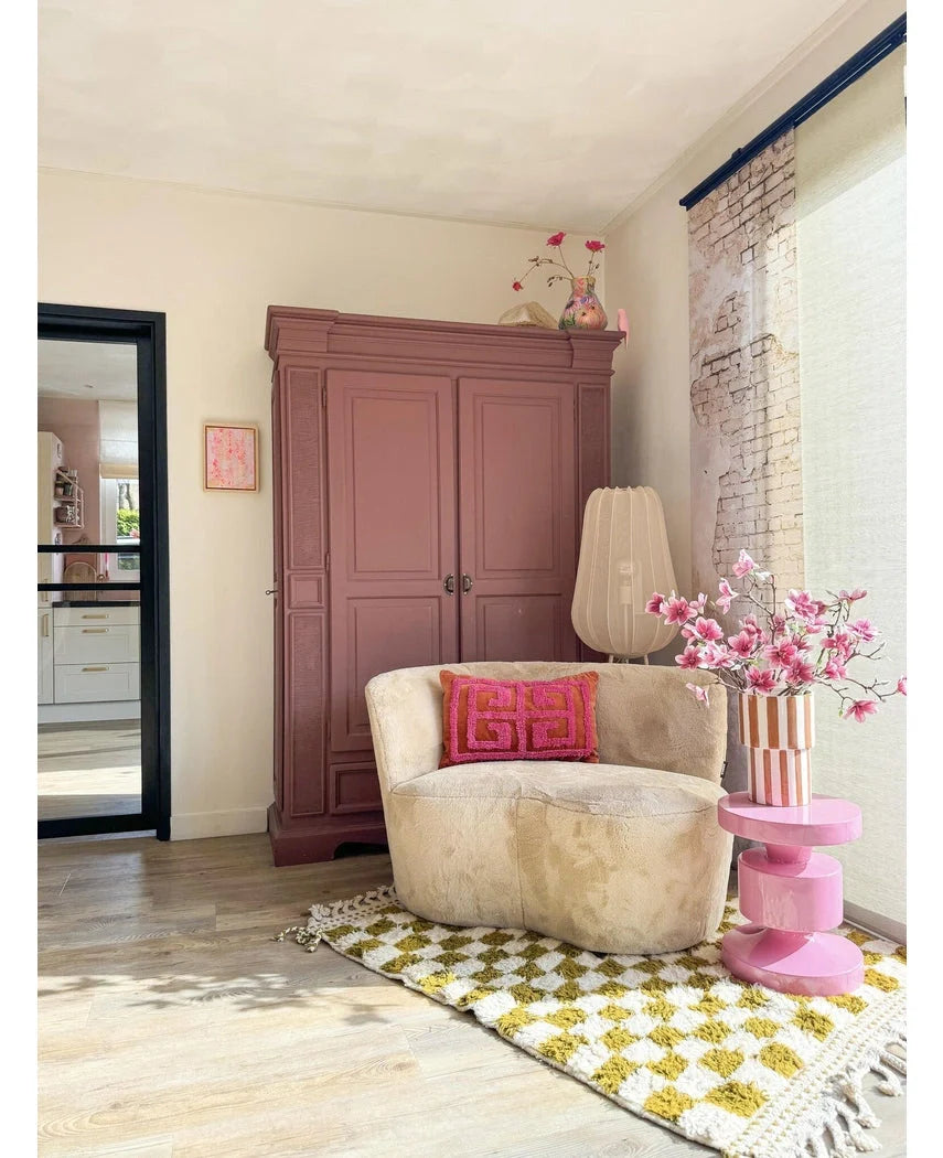 En hyggelig læsekrog har en cremefarvet stol, lyserøde puder, et gult mønstret tæppe og en lilla garderobe. The Artificial Branch Magnolia lilleblomstret 89 cm pink står på et pink sidebord, mens store vinduer fylder dette indbydende rum med blødt lys.